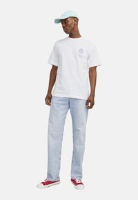 Camiseta blanca de algodón con logo y gráfico azul, combinada con jeans de mezclilla azul claro. Zapatillas rojas con acentos blancos y una gorra azul claro.