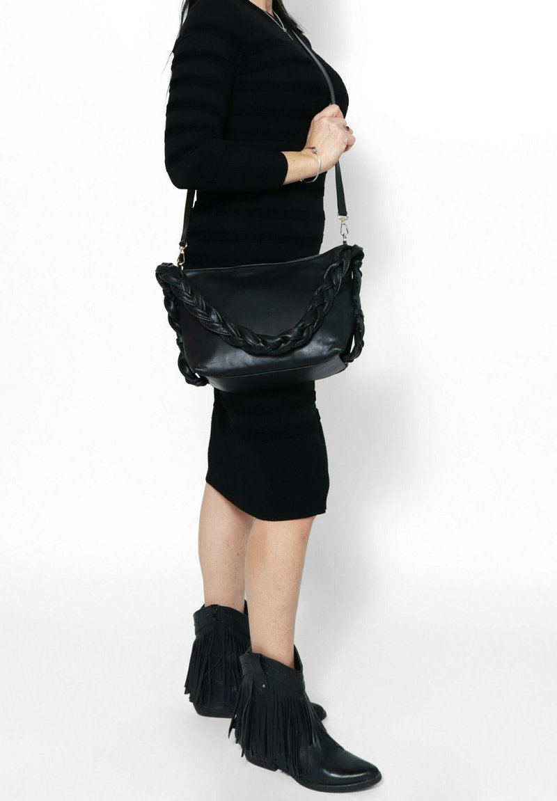 Zwarte leren schoudertas met een gevlochten handvat, een soepele textuur en gouden accenten. Gecombineerd met een aansluitende zwarte jurk en fringeboots in het zwart.