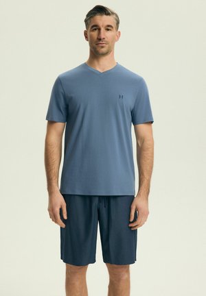 Mężczyzna stojący przodem, ubrany w gładką niebieską koszulkę z krótkim rękawem i dekoltem w serek oraz ciemnoniebieskie spodenki do kolan, na jasnym tle.