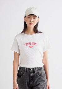 Camiseta blanca de algodón con bordado rojo de "TOMMY JEANS" en el pecho, combinada con jeans negros de cintura alta y una gorra de color claro.
