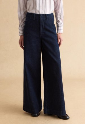 Mujer con pantalones vaqueros de tiro alto y pierna ancha de color azul oscuro, combinados con una camisa blanca abotonada con finas rayas verticales.