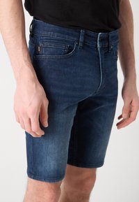 Shorts di jeans blu scuro con finitura sbiadita, dotati di cinque tasche, passanti per cintura e chiusura a bottone. Taglio aderente sopra il ginocchio.