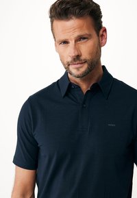 Mörkblå pikétröja i smidigt bomull med krage, korta ärmar och en diskret logotyp på vänster bröst. Klassisk passform och design.