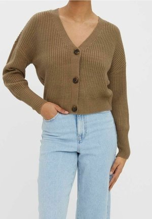 Vrouw draagt een bruine gebreide vest met knopen, een V-hals en lichtblauwe hoge taille jeans, hand in de zak tegen een effen achtergrond.