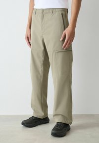 Bēšas kravas bikses ar rāvējslēdzēja kabatām, elastīgu jostasvietu un taisnām kājām. Izgatavotas no viegla, izturīga auduma.