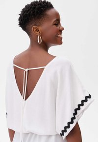 Witte top met een V-hals aan de achterkant, met korte mouwen en zwarte scalloped accenten en een tailleband. Gemaakt van lichtgewicht, gestructureerde stof.