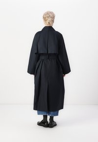 Dlouhý černý trenčkot s páskem v pase, volnými rukávy a zkráceným zadním designem. Hladká tkanina s klasickým siluetou.