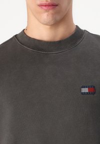 Tmavošedý bavlnený sveter s rebrovaným okrúhlym výstrihom a malou obdĺžnikovou nášivkou "Tommy Jeans" v červenej, bielej a modrej farbe na hrudi.