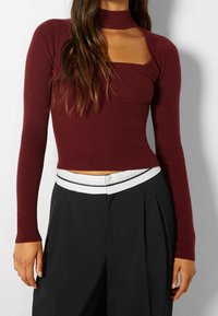 Top de manga larga de canalé en color burdeos con escote cuadrado y cuello alto, combinado con pantalones negros que tienen un acento de cintura blanca.