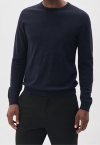 Granatowy sweter z dzianiny z okrągłym dekoltem i prążkowanymi mankietami. Materiał jest miękki o gładkiej fakturze, noszony na czarnych dopasowanych spodniach.