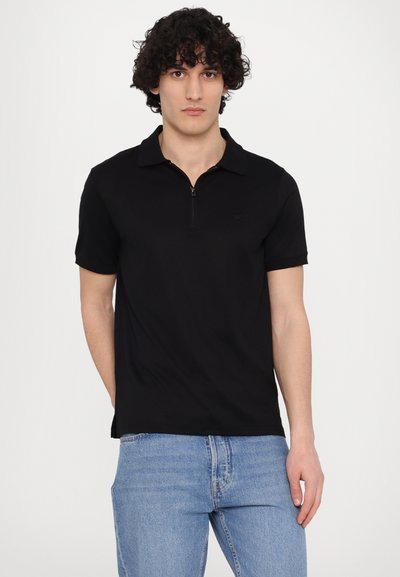 Μαύρο πόλο μ�πλουζάκι με κλείσιμο στο γιακά, κοντά μανίκια και διακριτό λογότυπο στο στήθος. Συνδυασμένο με ανοιχτό μπλε τζιν.