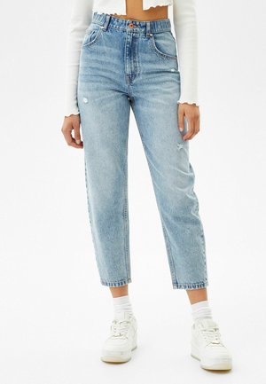 Person trägt hellblaue hoch taillierte Cropped-Jeans mit leichter Used-Optik, weiße Sneakers, weiße Socken und ein weißes langärmliges Cropped-Top.