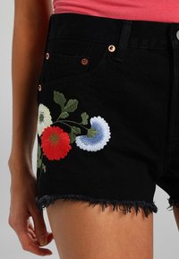 Pantalones cortos de mezclilla negra con dobladillo deshilachado, con bordados florales coloridos en rojo, azul y blanco en el lado izquierdo. Accesorios en tono cobre.