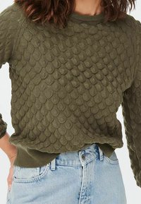 Maglione lavorato a maglia verde oliva con un motivo a scaglie testurizzato, scollatura rotonda e polsini e orlo a coste. Abbinato a denim azzurro chiaro.