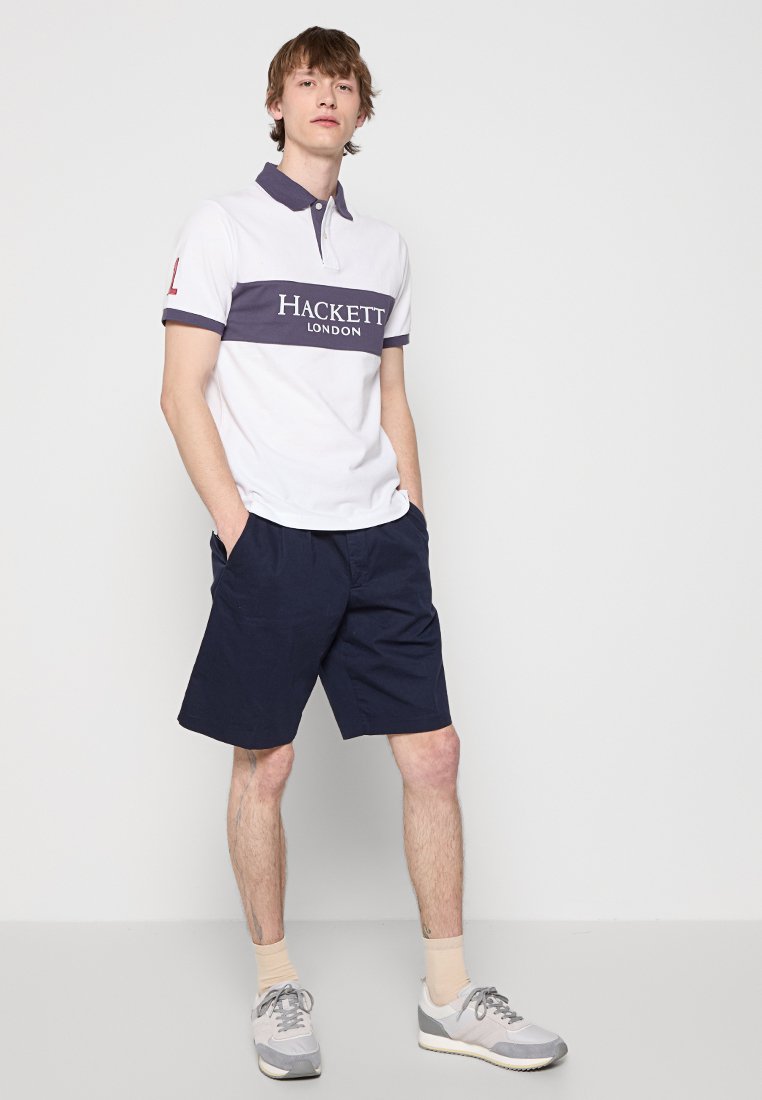 Jeune homme debout avec les mains dans les poches, portant un polo blanc et marine de Hackett London, un short marine, des chaussettes beiges et des baskets grises contre un mur blanc.