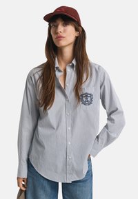 Donna con un berretto bordeaux, camicia a righe blu con bottoni e un ricamo sulla tasca, e jeans blu, in piedi con la mano sui fianchi.