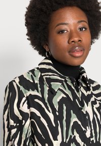 Camisa de botão com um padrão abstrato em preto, verde e bege, com gola e botões pretos, feita de um tecido texturizado.