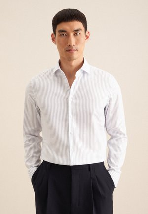 Camicia a righe bianche con linee verticali blu, maniche lunghe, collo button-down e una texture di tessuto liscia, indossata con pantaloni scuri.