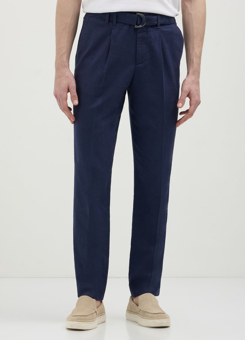 Pantalons en lin bleu marine avec une coupe décontractée, deux plis à l'avant, une ceinture à boucle argentée et des poches latérales. Portés avec des chaussures beiges à enfiler.