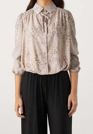 Femme portant un chemisier beige clair à motifs avec des boutons et un col noué, associé à un pantalon noir plissé taille haute.