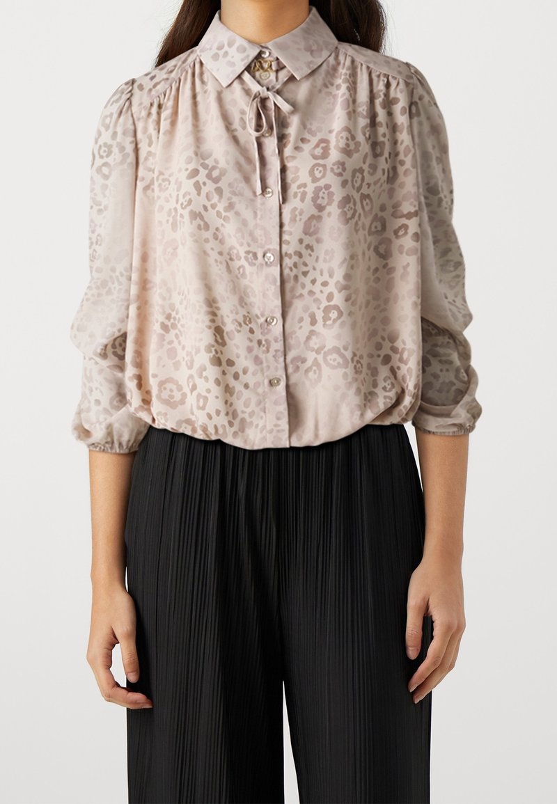 Femme portant un chemisier beige clair à motifs avec des boutons et un col noué, associé à un pantalon noir plissé taille haute.