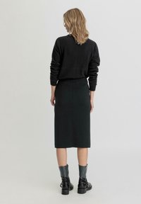 Mustat neuleet, joissa on pitkät hihat, yhdistettynä tummanvihreään kynähameeseen. Mallilla on jalassaan mustat nilkkurit, joissa on paksu korko.