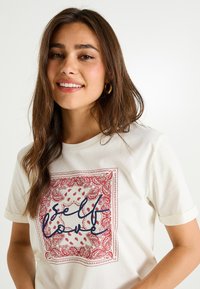 Camiseta de algodón blanca con un cuadrado de patrón paisley rojo y el texto "autoamor" en azul oscuro. Mangas cortas con puños enrollados.