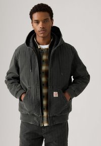 Dark gray hooded jacket with vertical stripes, front pockets, and ribbed cuffs. Underneath, a plaid button-up shirt in earth tones.