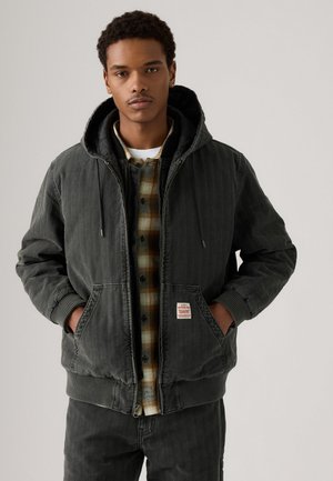 Dark gray hooded jacket with vertical stripes, front pockets, and ribbed cuffs. Underneath, a plaid button-up shirt in earth tones.