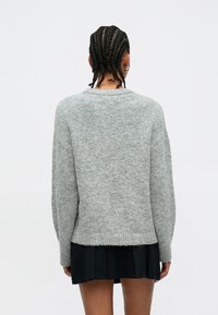 Grau gestrickter Pullover mit lockerem Schnitt, geripptem Saum und heruntergesetzten Schultern. Weiche Textur, mittellang, kombiniert mit einem plissierten schwarzen Rock.