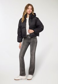 Giacca a piumino nera con cappuccio, design corto e texture trapuntata, abbinata a jeans grigi a vita alta e sneaker bianche.