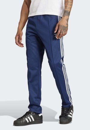 Muž oblečený v modrých teplácích Adidas s bílými pruhy po stranách, černými teniskami Adidas a bílém tričku stojí s uvolněnýma rukama.