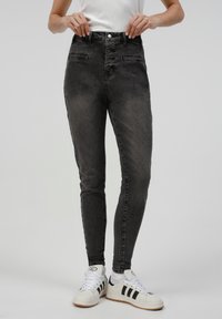 Jeans ajustados negros con una textura desgastada, cintura alta, bolsillos frontales y una cremallera con botones. Combinados con zapatillas blancas con rayas negras.
