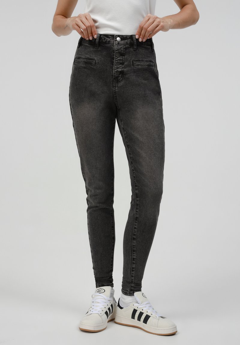 Jeans ajustados negros con una textura desgastada, cintura alta, bolsillos frontales y una cremallera con botones. Combinados con zapatillas blancas con rayas negras.
