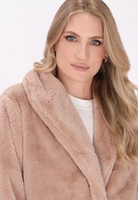 Rochie din blană sintetică de culoare bej deschis, cu guler tip șal, textură moale și deschidere frontală. Pur tată peste un top alb, are o finisare netedă.