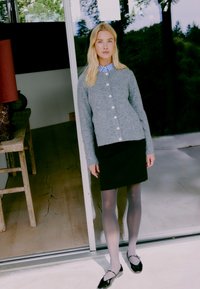 Grau gestrickte Strickjacke mit Knöpfen, kombiniert mit einem schwarzen Rock, durchsichtigen grauen Strumpfhosen und schwarzen Ballerinas. Innenraum mit Holzboden.