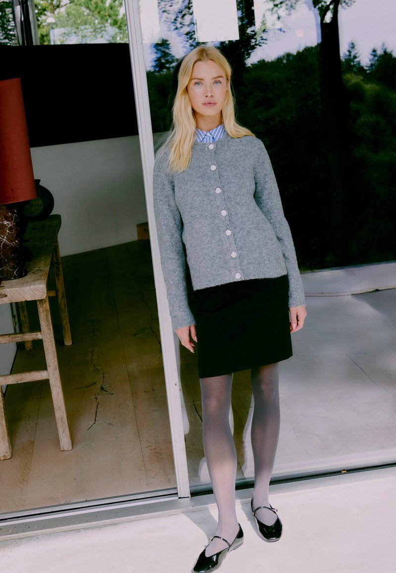 Grau gestrickte Strickjacke mit Knöpfen, kombiniert mit einem schwarzen Rock, durchsichtigen grauen Strumpfhosen und schwarzen Ballerinas. Innenraum mit Holzboden.