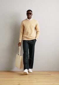 Sudadera beige, pantalones negros, zapatillas blancas y una bolsa de lona. Las gafas de sol y una camisa de cuello completan el look. Texturas suaves en general.