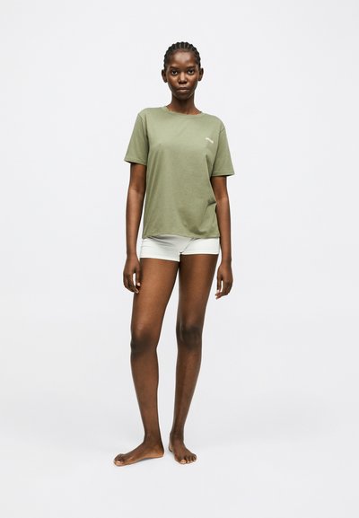 Jeune femme aux cheveux tressés, debout pieds nus, portant un T-shirt à manches courtes vert olive et un short blanc, posant devant un fond uni.