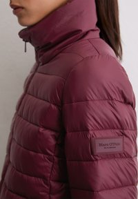 Bordeaux pufferjack met een hoge kraag, geborduurd textuur en een stoffen label op de mouw met de tekst "Marc O'Polo." Gladde afwerking.