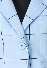 Lys blå blazer med rutete mønster i hvitt og svart. Har spiss krage og en lys blå knapp. Glatt tekstur.