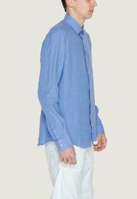 Uomo in camicia a maniche lunghe blu e pantaloni bianchi, in profilo contro uno sfondo chiaro e semplice.