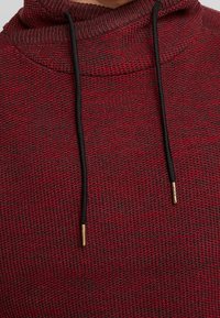 Bordeauxroter Strick-Hoodie mit hohem Kragen und schwarzen Kordeln, die goldene Metallenden und eine strukturierte Stoffoberfläche aufweisen.