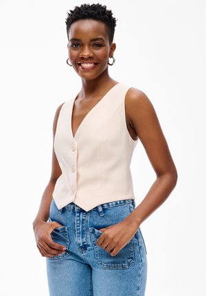 Uśmiechnięta kobieta w kolczykach-kołach, bezrękawniku w beżowym kolorze zapinanym na guziki oraz wysokich niebieskich dżinsach z rękami w przednich kieszeniach.