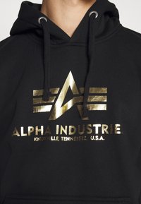 Felpa nera con cappuccio caratterizzata da un logo dorato metallico con design geometrico e testo "ALPHA INDUSTRIE" e dettagli sulla posizione. Cappuccio regolabile con cordino.