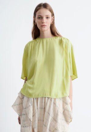 Young woman wearing a loose lime green top and beige plaid skirt with ruffles, standing against a plain light background.