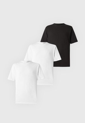 Jeune homme debout portant un t-shirt blanc uni à manches courtes, un pantalon noir et des baskets beiges sur fond blanc.
