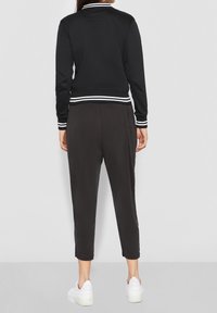 Donna che indossa pantaloni cropped neri, giacca nera con strisce bianche sui polsini e sull'orlo, e sneakers bianche, in piedi contro uno sfondo neutro.