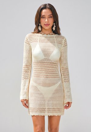 Donna che indossa un vestito in maglia all'uncinetto color crema sopra un bikini, in piedi su uno sfondo chiaro e semplice, con orecchini lunghi e capelli sciolti.