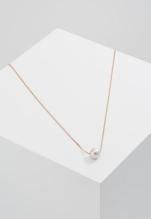 Collier délicat en chaîne d'or avec un pendentif unique en perle blanche ronde reposant sur une surface géométrique blanche.